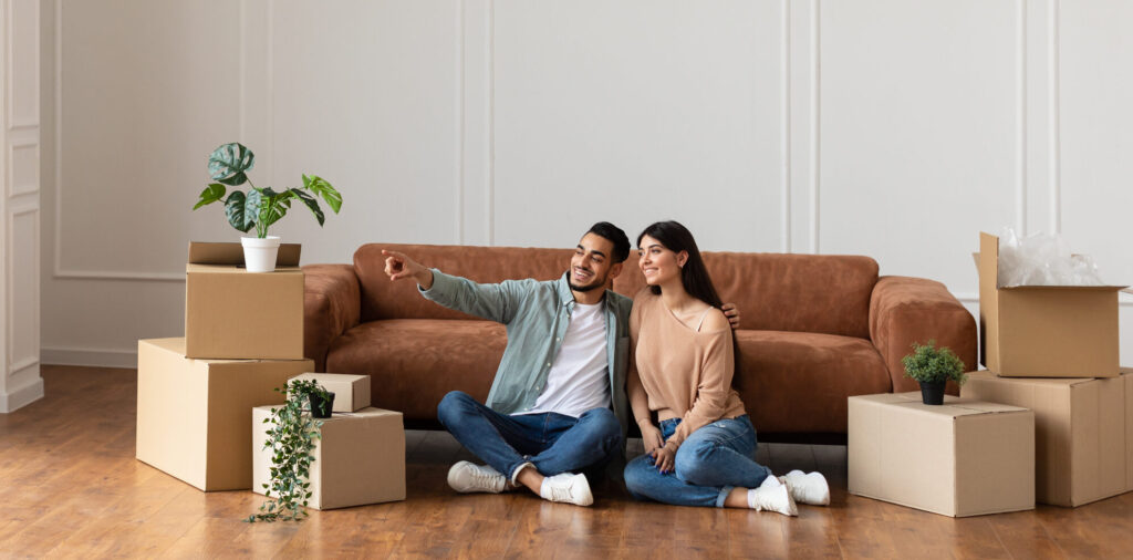 Happy man and sitting on floor choosing interior design Portrait of smiling family sitting on the floor leaning on couch in new empty apartment. Guy pointing finger aside at free space embracing wife, choosing wall color and interior design repair, banner